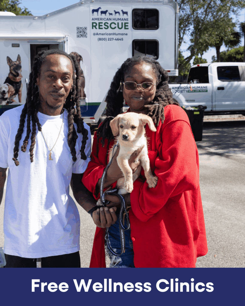 A couple with their puppy at a free wellness clinic. Pictured in the background is a white trailer with the words American Humane Rescue.